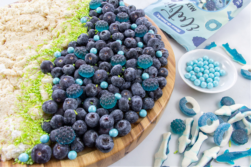 ocean themed grazing board being made with the ocean made up of blueberries, blue chocolate balls, blue gummy candy, and blue gummy sharks. 