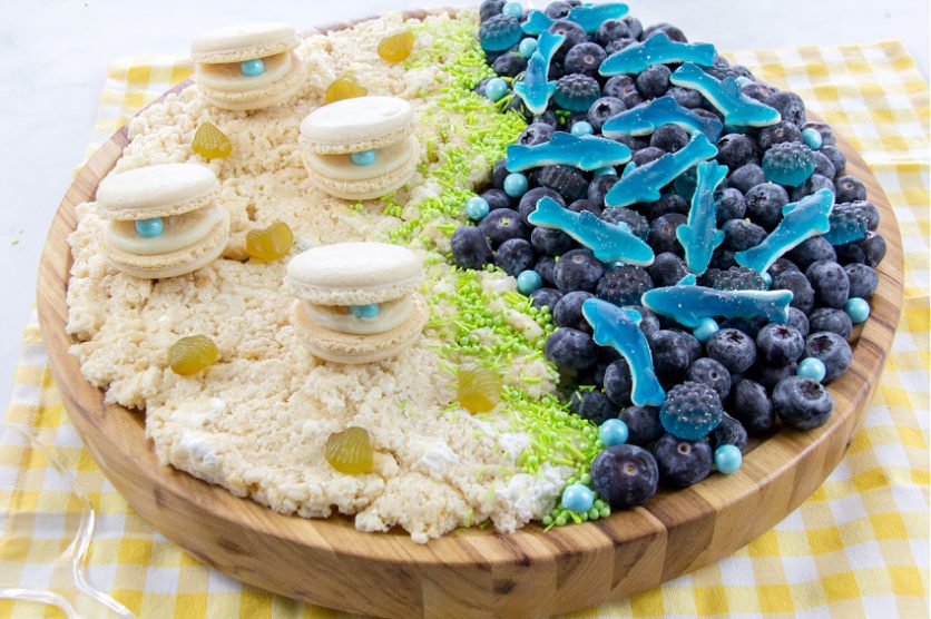 ocean themed charcuterie board with rice krispies sand, macaron clams, blueberry and gummy ocean with gummy sharks