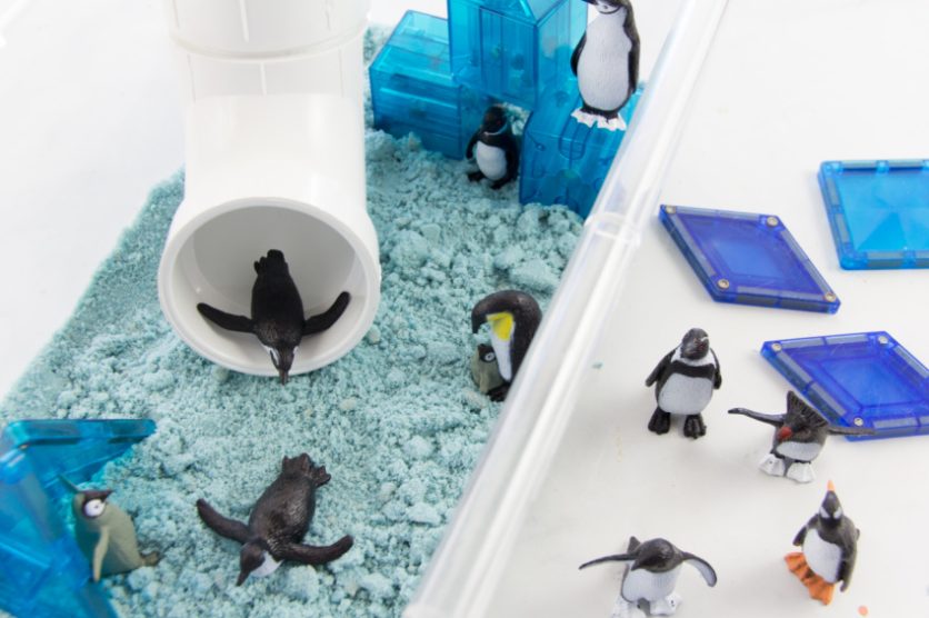 winter sensory bin with a tube slide, magnetic blocks, blue sand, and penguins.