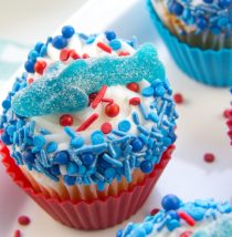 cupcakes decorated with blue and red sprinkles and topped with a gummy shark for shark week