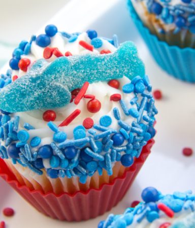 cupcakes decorated with blue and red sprinkles and topped with a gummy shark for shark week