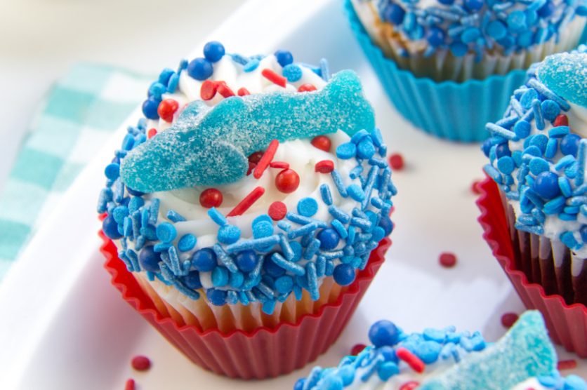 cupcakes decorated with blue and red sprinkles and topped with a gummy shark for shark week