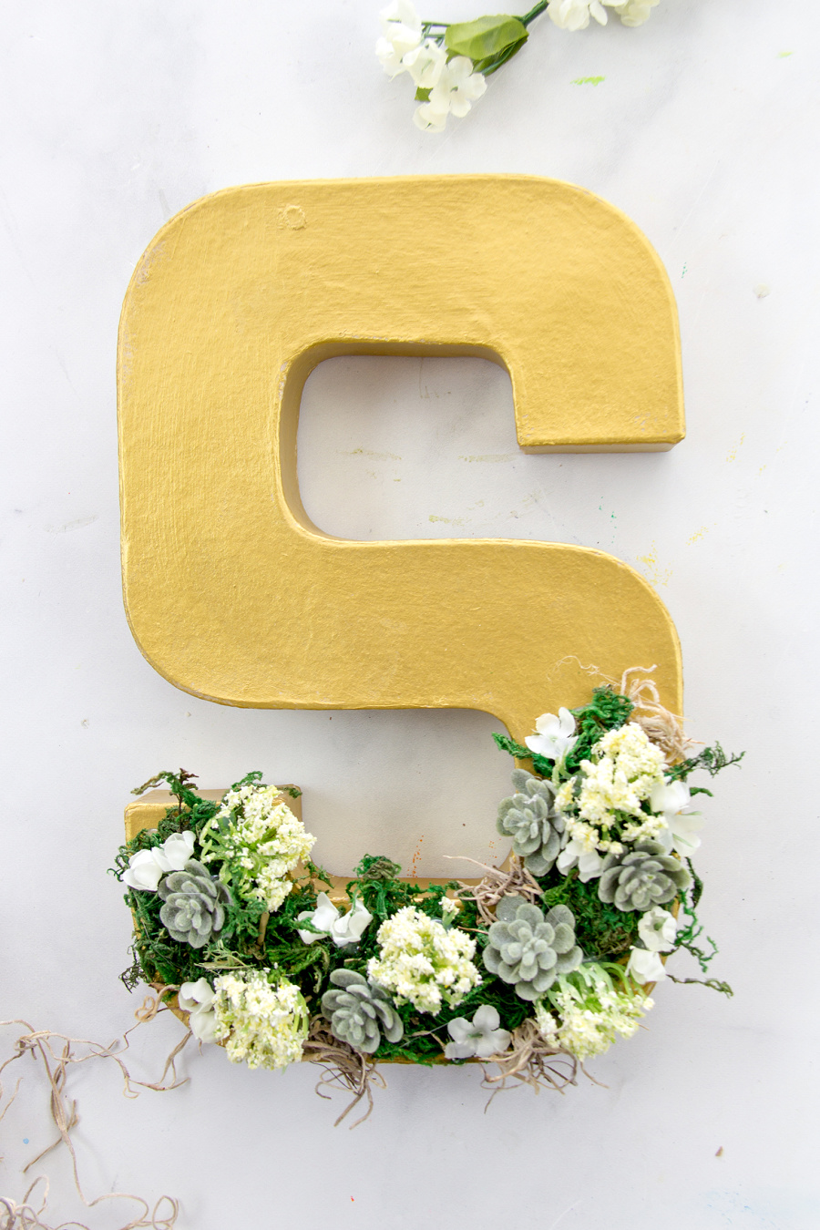 A paper mache letter S painted with gold paint and decorated with faux succulents, moss, and flowers.