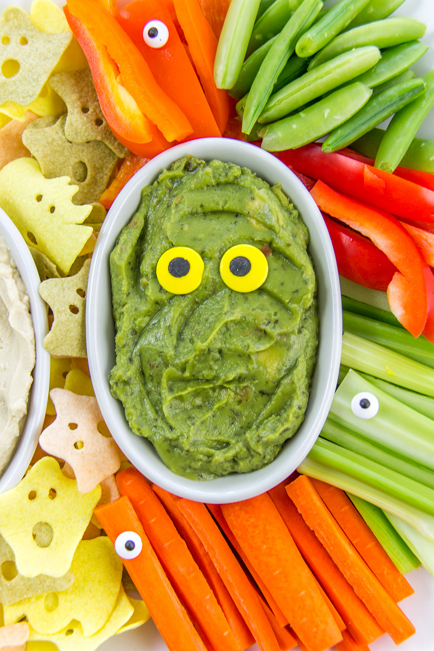 guacamole in a bowl with yellow candy eyeballs surround by halloween veggie chips and vegetables for dipping