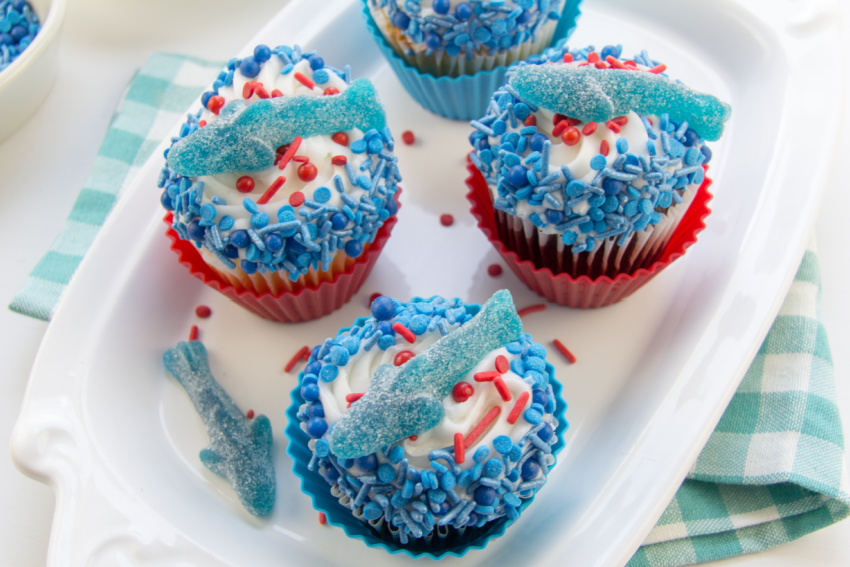 cupcakes decorated in blue and red sprinkles and topped with a gummy shark for shark week