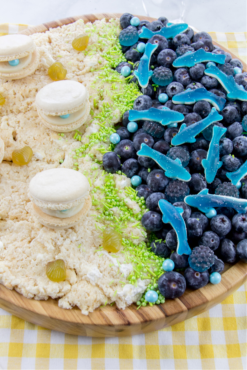 ocean themed party food grazing board with rice krispie treats, macaron clams, blueberries, and gummy sharks. 